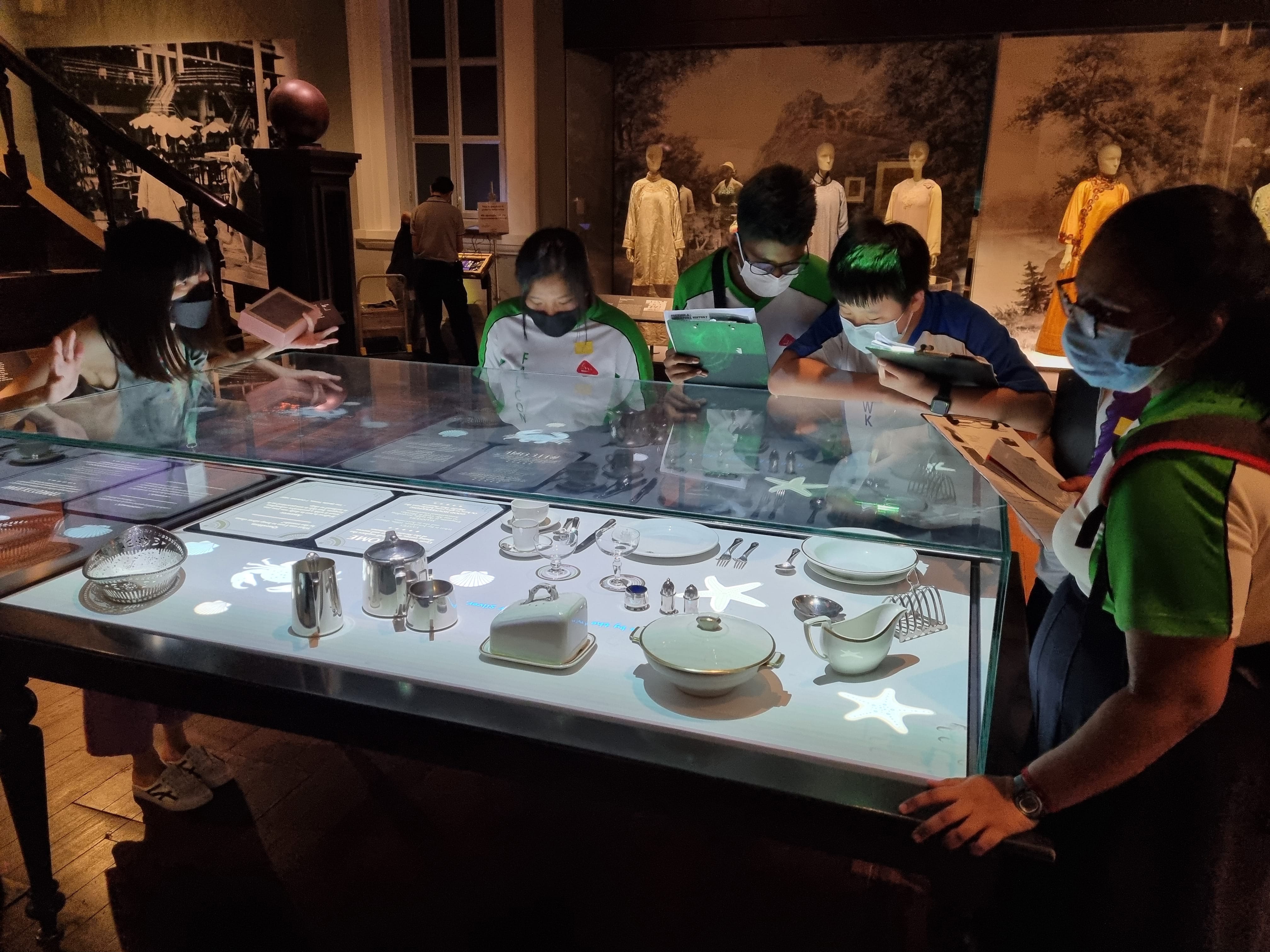 Students in masks examine a lit glass display table of dishes and silverware in a museum gallery.